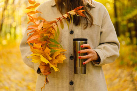 A woman is holding a thermo cup and a bouquet of autumn leaves. Autumn outdoor activity.