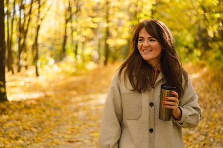 A young woman in a long shirt with a thermo mug in her hands walks through the autumn forest.