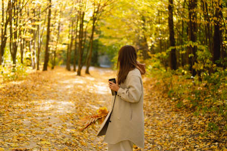 A young woman in a long shirt with a thermo mug in her hands walks through the autumn forest.の写真素材