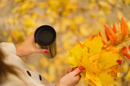 A woman is holding a thermo cup and a bouquet of autumn leaves. Autumn outdoor activity.