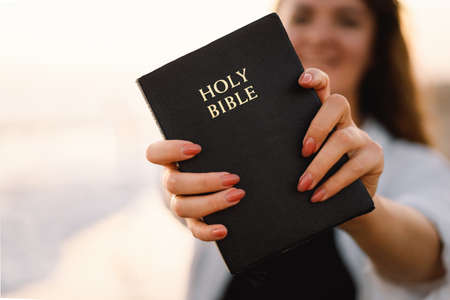 Christian woman holds bible in her hands. Reading the Holy Bible on the sea during beautiful sunsetの写真素材
