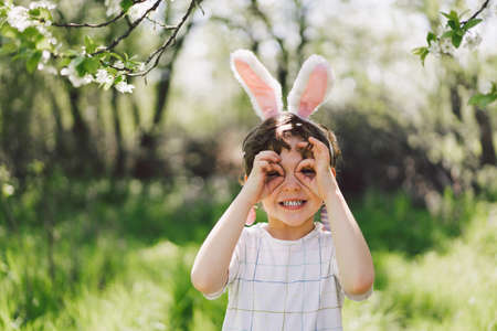 Funny boy with eggs basket and bunny ears on Easter egg hunt in sunny spring gardenの写真素材