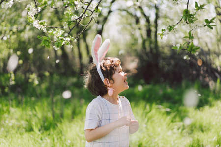 Funny boy with eggs basket and bunny ears on Easter egg hunt in sunny spring gardenの写真素材
