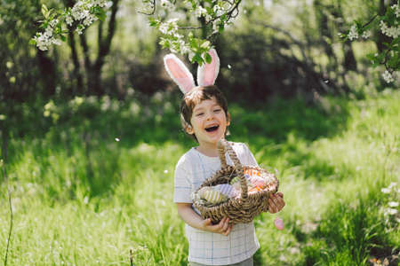 Funny boy with eggs basket and bunny ears on Easter egg hunt in sunny spring garden