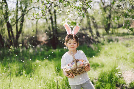Funny boy with eggs basket and bunny ears on Easter egg hunt in sunny spring garden