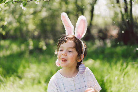 Funny boy with eggs basket and bunny ears on Easter egg hunt in sunny spring gardenの写真素材