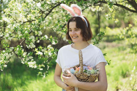 Funny teen girl with eggs basket and bunny ears on Easter egg hunt in sunny spring garden