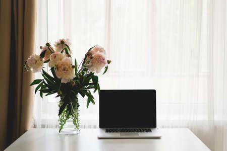 Laptop and vase with bouquet white pink peonies flowers on a table.の写真素材