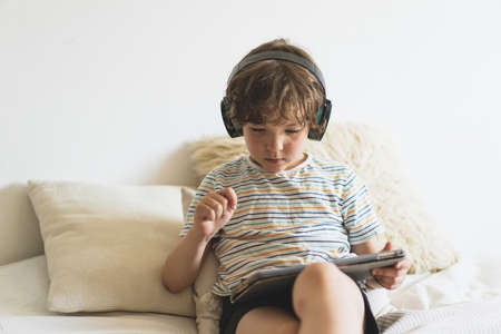 Cute boy lying on the bed at home and playing with pc tablet with wireless headphones