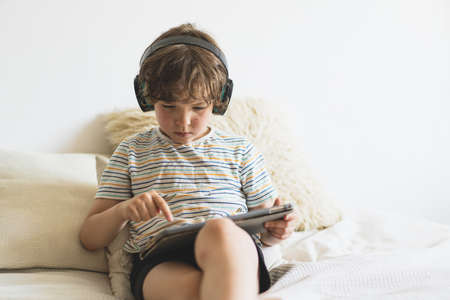 Cute boy lying on the bed at home and playing with pc tablet with wireless headphonesの写真素材