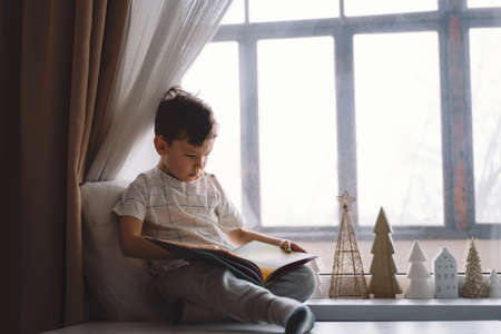 Cute boy is reading a book sitting on the windowsill near the window. Cozy home