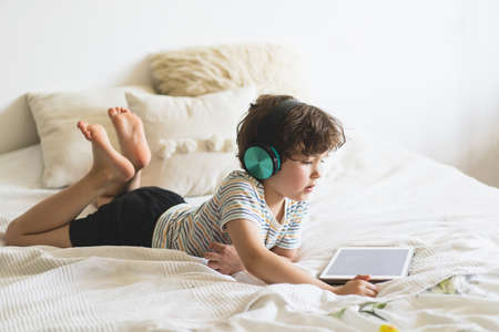 Cute boy lying on the bed at home and playing with pc tablet with wireless headphonesの写真素材