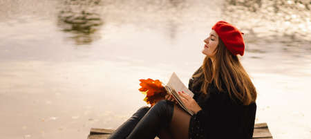 Woman in a red beret reading book on wooden pontoon. Autumn season.の写真素材