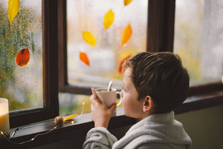 Boy wrapped in a blanket drink hot tea and looks out the open window at the wonderful autumn nature