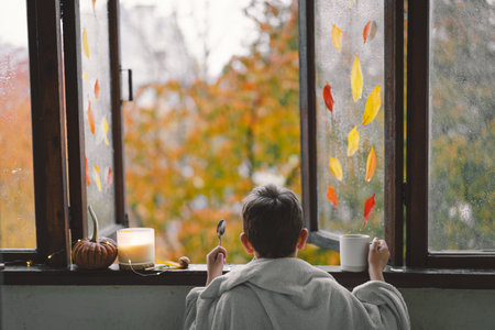 Boy wrapped in a blanket drink hot tea and looks out the open window at the wonderful autumn nature