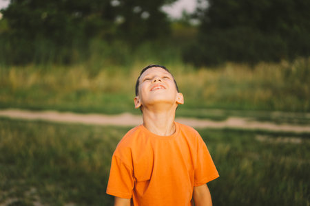 Portrait of a smiling little boy in a orange T-shirt and playing outdoors on the field at sunsetの写真素材