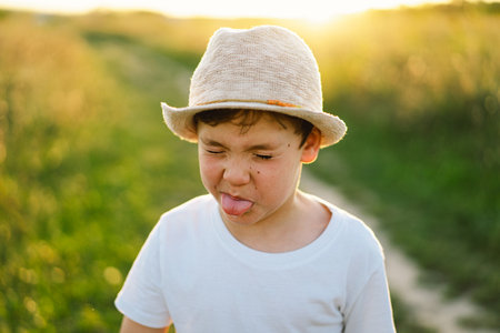 Portrait of a smiling little boy in a white T-shirt and hat playing outdoors on the field at sunsetの写真素材