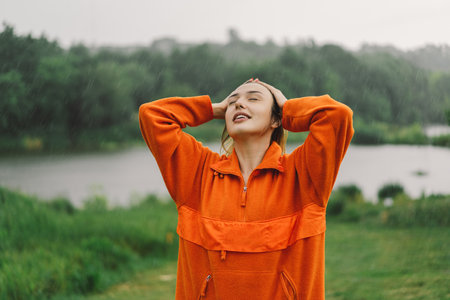 A young girl in an orange sweater is enjoying the rain on a summer day.