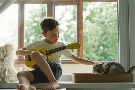 Cute boy learns to play the Ukulele guitar on the windowsill near the windowの写真素材