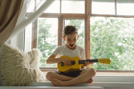 Cute boy learns to play the Ukulele guitar on the windowsill near the windowの写真素材