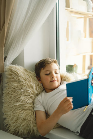 Cute boy reading a book on the windowsill near the window.の写真素材