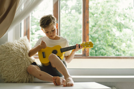 Cute boy learns to play the Ukulele guitar on the windowsill near the windowの写真素材