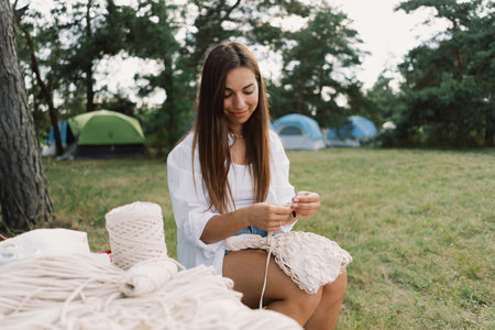Woman knits bag using macrame technique outdoors near tents. Outdoor hobbies.