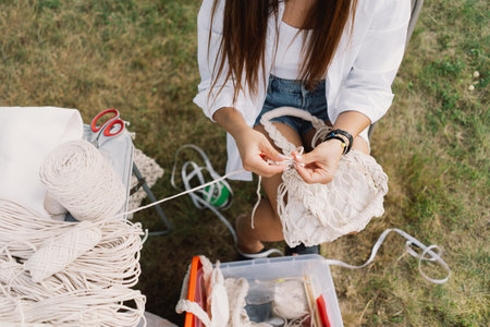 Woman knits bag using macrame technique outdoors near tents. Outdoor hobbies.