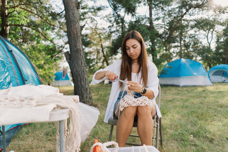 Woman knits bag using macrame technique outdoors near tents. Outdoor hobbies.