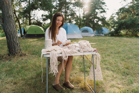 Woman knits bag using macrame technique outdoors near tents. Outdoor hobbies.