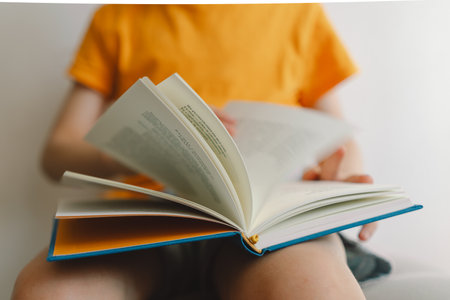 A cute boy wearing an orange T-shirt is sitting on a soft ottoman reading a book