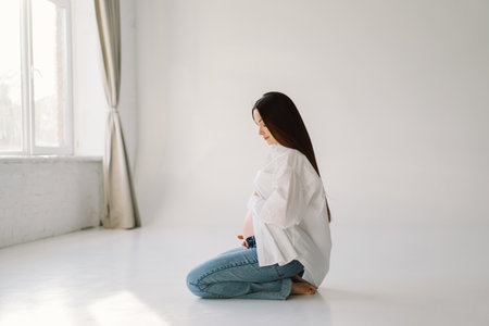 Cute young Pregnant girl sitting on the white studio in warm lightの写真素材
