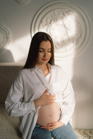 Cute young Pregnant girl sitting on the white studio in warm lightの写真素材