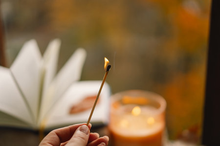 Hand with burning match lighting a candle on the windowsill with autumn decorの写真素材