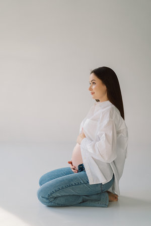 Cute young Pregnant girl sitting on the white studio in warm lightの写真素材