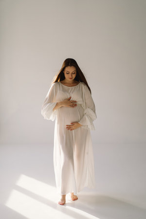 Cute young Pregnant girl sitting on the white studio in warm lightの写真素材