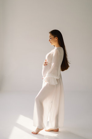 Cute young Pregnant girl sitting on the white studio in warm lightの写真素材