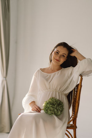 Cute young Pregnant girl sitting on the white studio in warm lightの写真素材