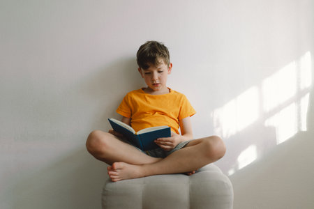 A cute boy wearing an orange T-shirt is sitting on a soft ottoman reading a bookの写真素材