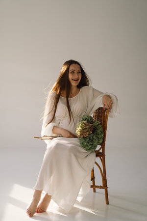 Cute young Pregnant girl sitting on the white studio in warm lightの写真素材