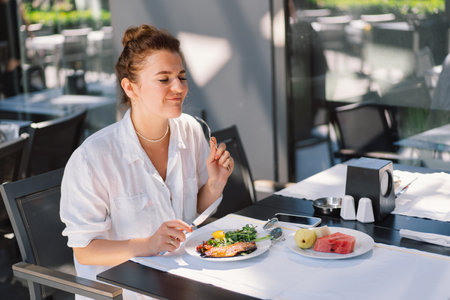 A woman in a white shirt eats lunch or breakfast outdoors in a cafe.の写真素材