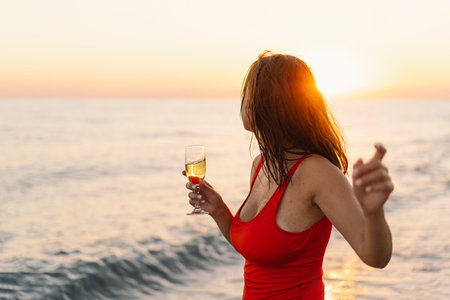 Smiling Woman in Red swimsuit Holding Wine Glass by the Sea at Sunsetの写真素材