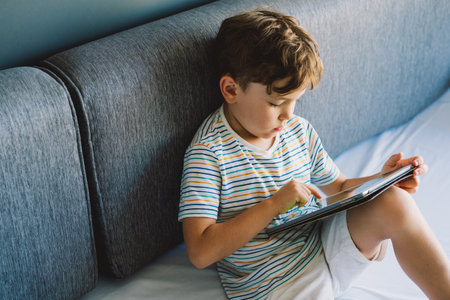 Young Boy Engrossed in playing tablet on a Cozy Couch During Afternoon Hours