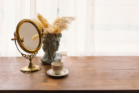 Elegant Vintage Mirror and Bust Vase With Pampas Grass Next to a Coffee Cup on a Wooden Tableの写真素材