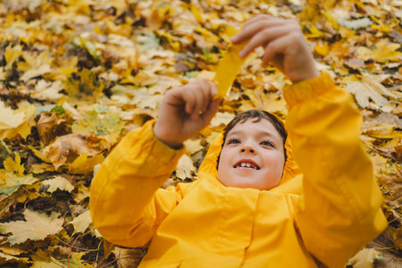 Joyful Child in Yellow Raincoat Laying in Autumn Leaves at a Parkの写真素材