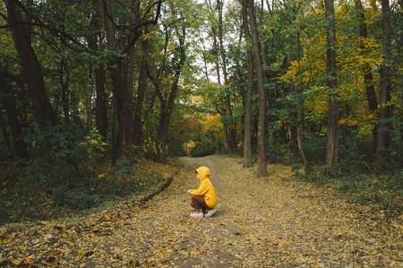 Child in Yellow Raincoat Walking Down Leaf-Covered Path in Autumn Forestの写真素材