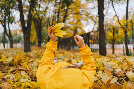 Joyful Child in Yellow Raincoat Laying in Autumn Leaves at a Parkの写真素材