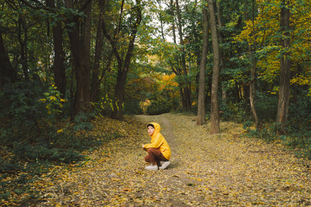 Child in Yellow Raincoat Walking Down Leaf-Covered Path in Autumn Forestの写真素材
