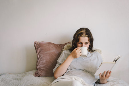Young woman reading a book while sipping coffee at homeの写真素材