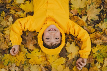 Joyful Child in Yellow Raincoat Laying in Autumn Leaves at a Parkの写真素材